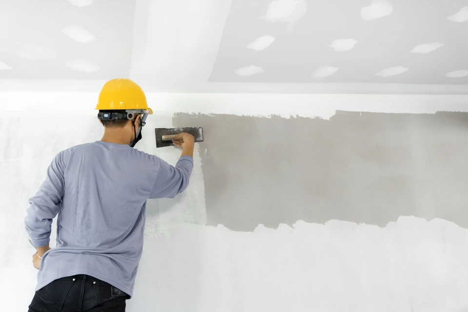 Photographe professionnelle en couleurs montrant l'intérieur d'un chantier de rénovation résidentielle avec plusieurs ouvriers en tenues professionnelles (combinaisons, casques) travaillant simultanément sur différents aspects : un électricien posant une prise murale, un menuisier ajustant une porte, un ouvrier inspectant une zone d'isolation, et un coordinateur ou contremaître consultable au centre de la scène, tablette ou documents en main, supervisant l'ensemble des travaux. La scène doit communiquer l'harmonie, la coordination et la gestion intégrée d'un chantier professionnel complet.