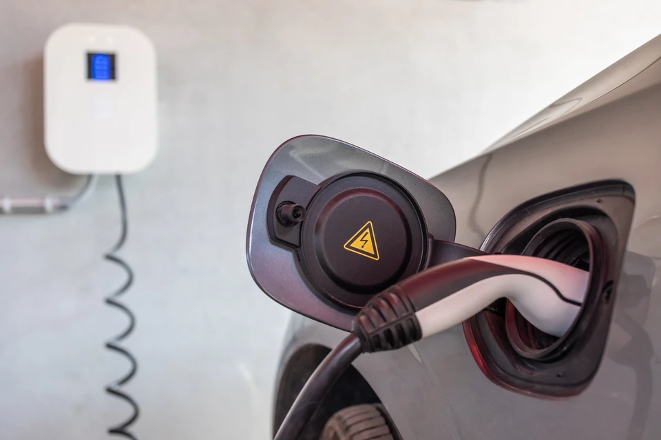 Une voiture électrique garée sur le parking ou l'allée d'une maison résidentielle, avec une borne de recharge murale moderne et élégante visible à proximité immédiate du véhicule. La borne est bien illuminée et clairement identifiable. L'arrière-plan montre une maison de style contemporain ou traditionnel. La scène doit transmettre la sécurité, la commodité et la simplicité de la recharge à domicile.